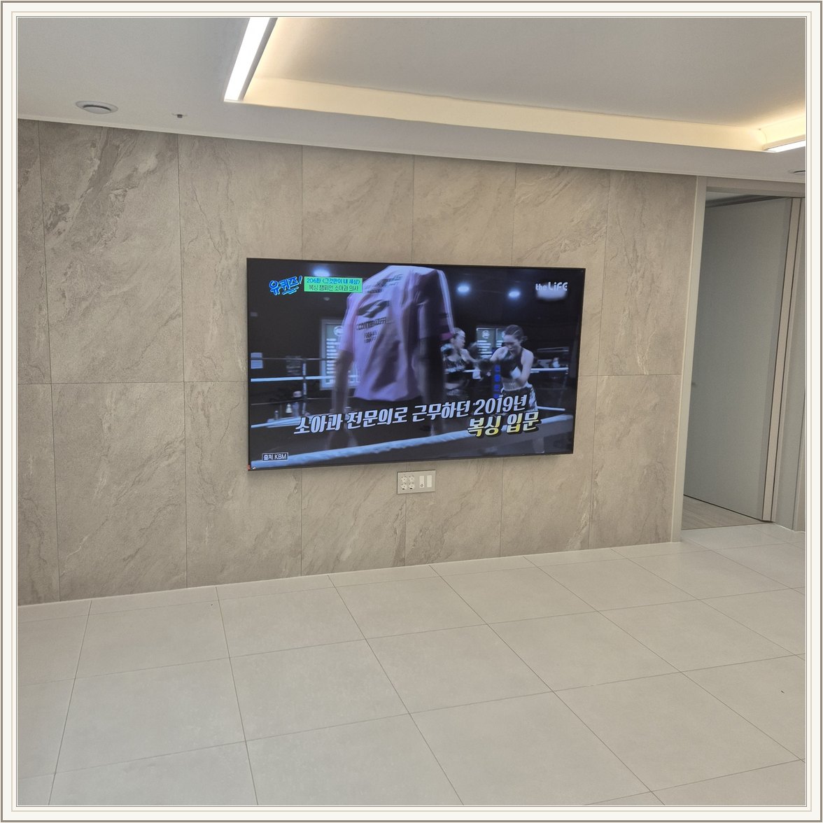 석재 느낌 벽면을 정면에서 본 TV / 넓은 바닥 여유와 조명 라인이 동시에 보입니다