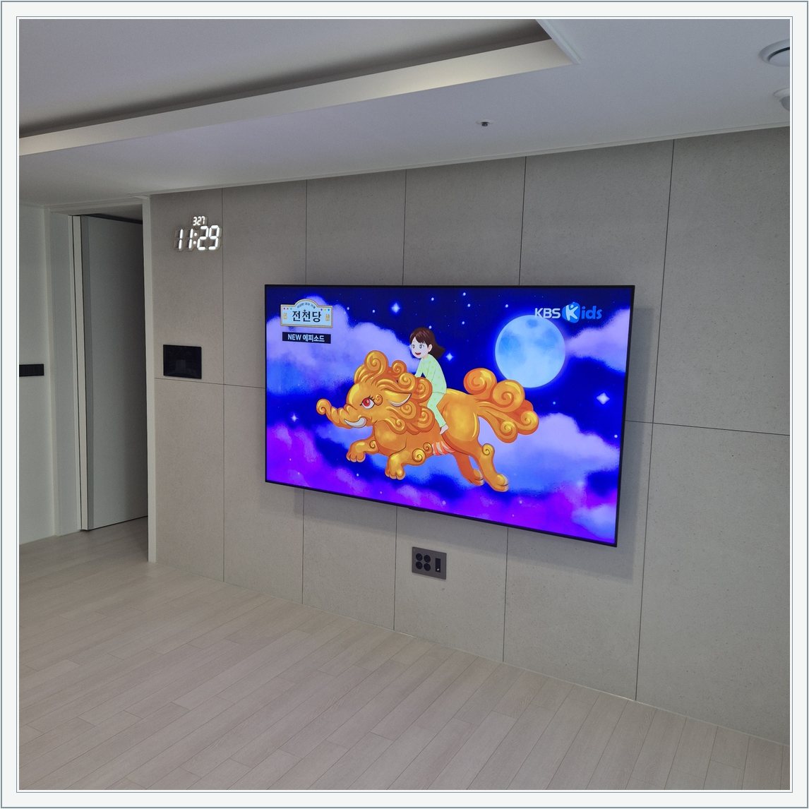 회색 판넬 벽 위의 화면과 디지털 시계 / 벽면 패널 이음선과 TV 중심의 배치를 비교할 수 있습니다