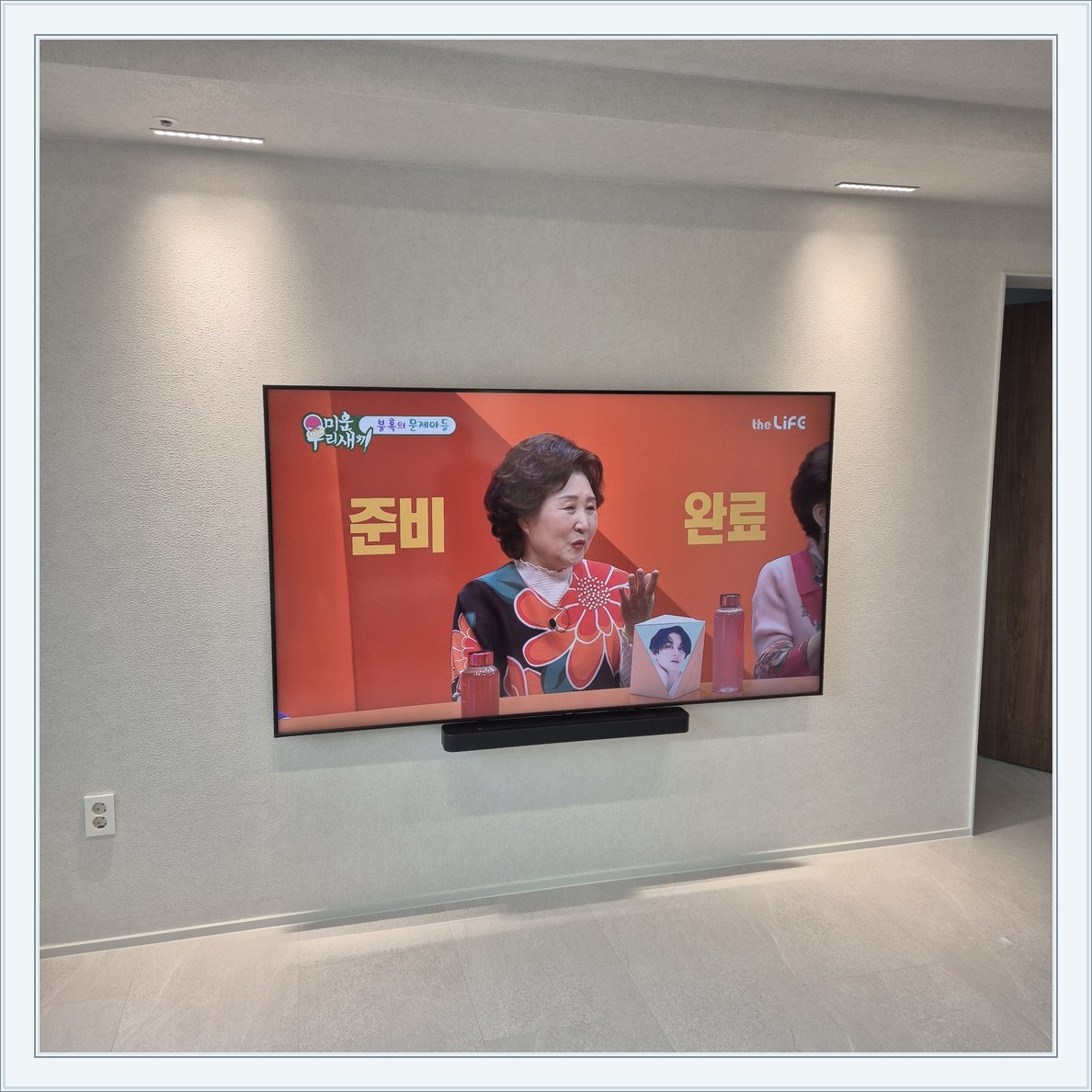 조명이 비추는 미색 벽과 사운드바 / 화면 아래 가로로 놓인 검은 음향기기가 선명합니다