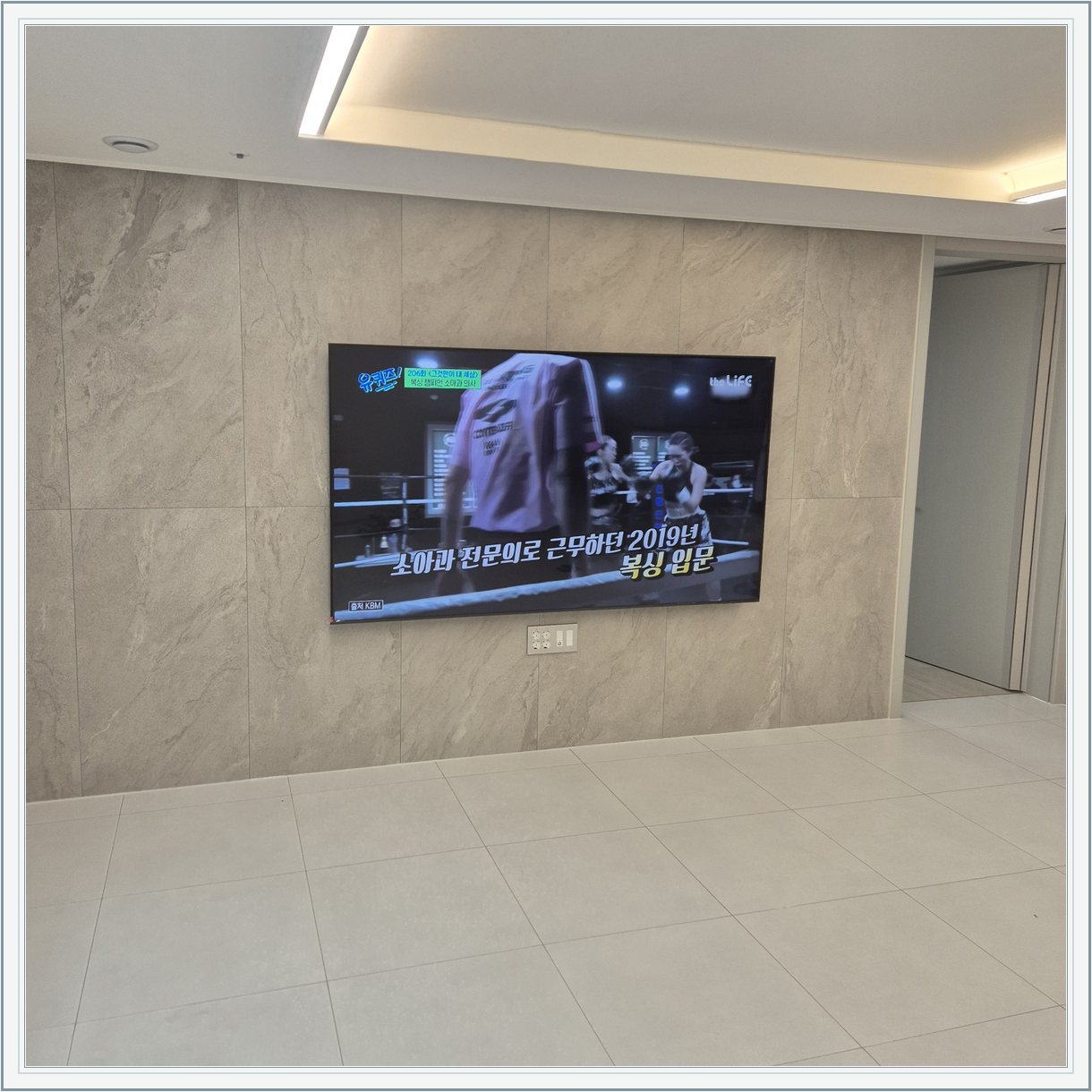 석재 느낌 벽면을 정면에서 본 TV / 넓은 바닥 여유와 조명 라인이 동시에 보입니다