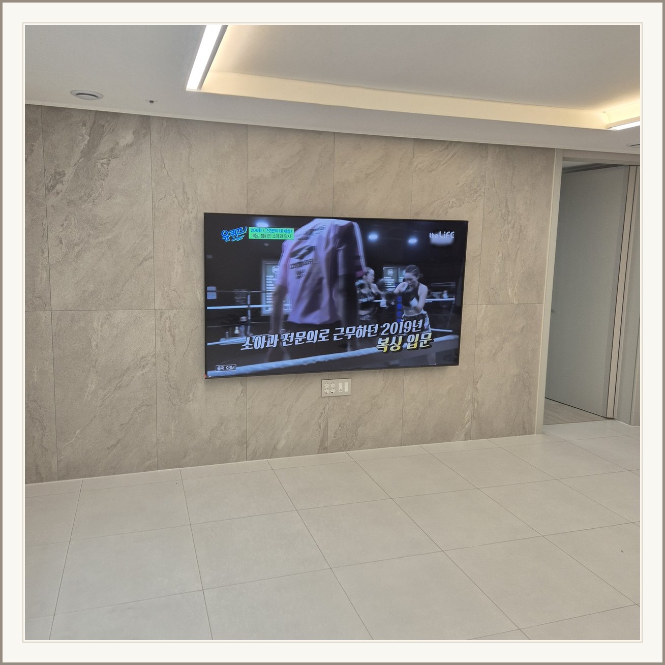 석재 느낌 벽면을 정면에서 본 TV / 넓은 바닥 여유와 조명 라인이 동시에 보입니다